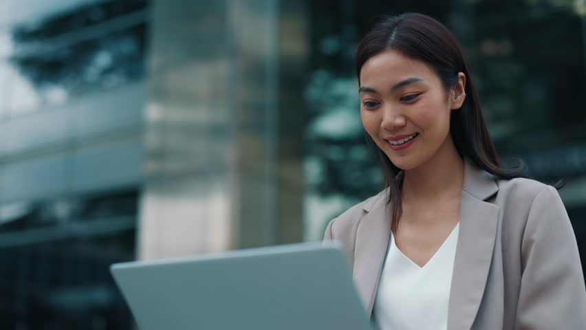 Portrait of Thai woman working on laptop sitting near office building. Young Asian female sends messages with cheerful smile sitting among urban environment. Positive lady entrepreneur - Powered by Shutterstock - Get 15% off with code: PIKWIZARD15