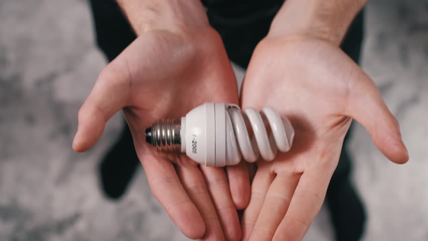 Close-up top view as male hands gently hold and present a spiral CFL energy-saving light bulb towards the camera. Zooming in slightly. Focus on energy conservation and modern lighting.