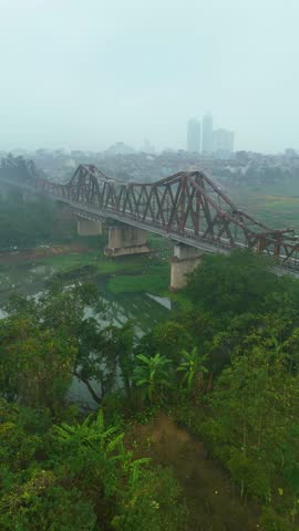 Vertical aerial view of Hanoi's Long Bien Bridge in Vietnam, with a hazy atmosphere caused by extreme pollution. The shot captures the iconic bridge stretching over the green landscape.