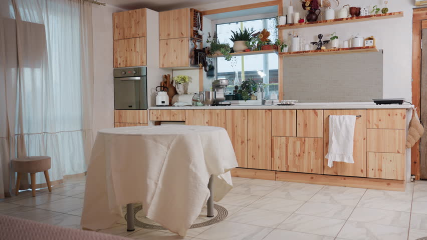 View of modern kitchen interior with wooden cabinets, countertop appliances, indoor plants, and cozy dining area with table covered in white cloth near large window