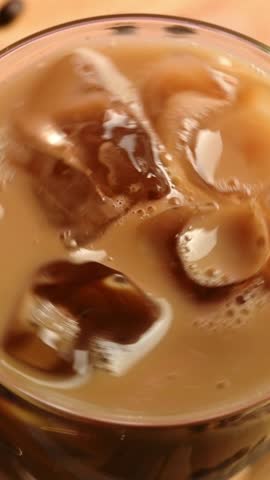 Ice cubes slowly rotate in iced coffee with milk inside a transparent glass, reflecting soft light and creamy brown tones in close-up view.