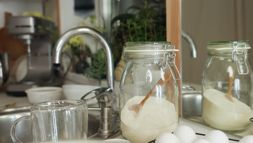 Hand view of chef picking up glass container filled with sugar from kitchen counter, clip top open, with background showing sink, plants, and natural kitchen setting in soft indoor lighting