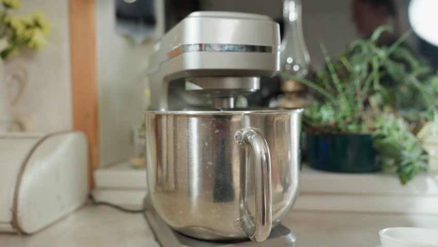 Chef turns off electric mixer after use in home kitchen as stainless bowl rests on mixer base, reflection of chef seen faintly in mirror, surrounded by plants and utensils in bright natural lighting
