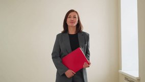 Confident businesswoman in gray suit holding red folder walking in office hallway with soft smile, ready for meeting or presentation, showing professionalism and corporate elegance - Powered by Shutterstock - Get 15% off with code: PIKWIZARD15