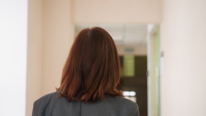 Close up back shot of woman walking purposefully down modern hallway, holding red folder, dressed in professional attire, exuding confidence and determination