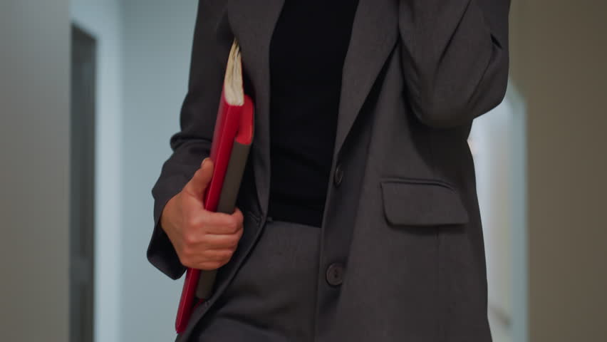 Close-up of businesswoman holding red folder in office corridor, dressed in gray suit, confident professional in modern work environment, career-focused, stylish business attire