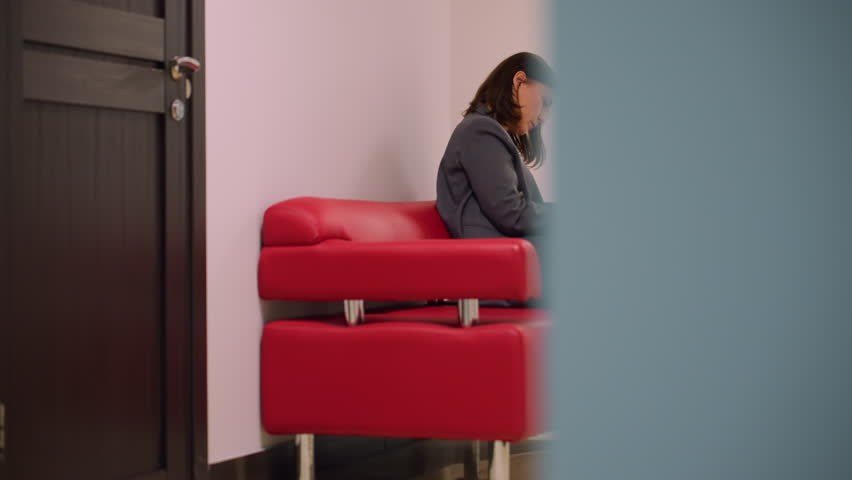 Woman in business attire sitting on red sofa in office hallway, focused on writing in notebook with pen. Calm, professional, modern workspace, preparing for meeting, taking notes