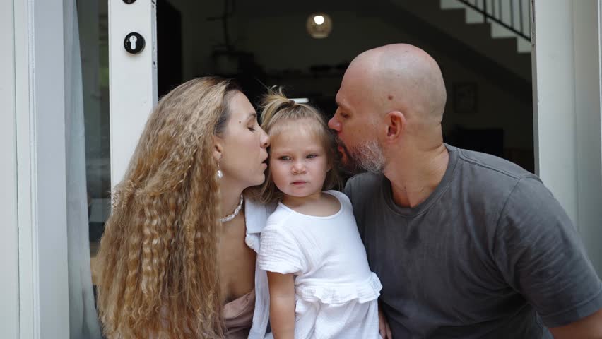 Loving parents kiss their daughter on cheeks, family of father, mother and child spend time together at home