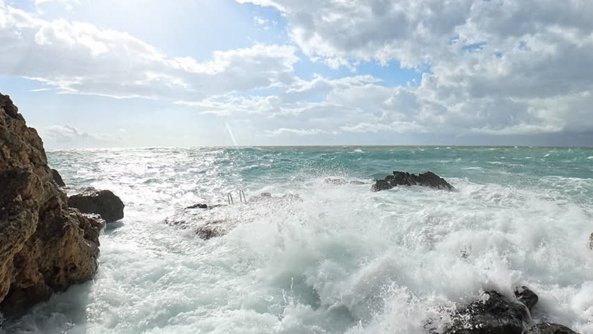 Powerful waves crashing over jagged coastal rocks, with foamy water spreading across the rugged terrain under a sunny sky filled with dramatic clouds