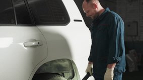 Mechanic bent with one hand on knee using torch light to inspect tire in auto repair workshop, blue pole in background, focused on vehicle maintenance - Powered by Shutterstock - Get 15% off with code: PIKWIZARD15