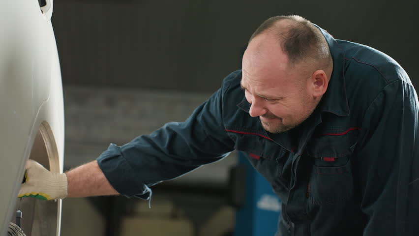 Mechanic in blue uniform using torchlight to inspect car tire in auto repair shop, focusing closely with blue lift in background, ensuring safety and maintenance in professional automotive workshop
