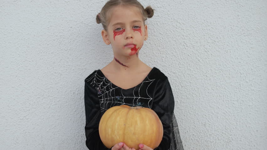 Girl in Halloween makeup holding pumpkin while standing against wall. She is dressed in a black costume with spiderweb details, and her face features festive makeup designs