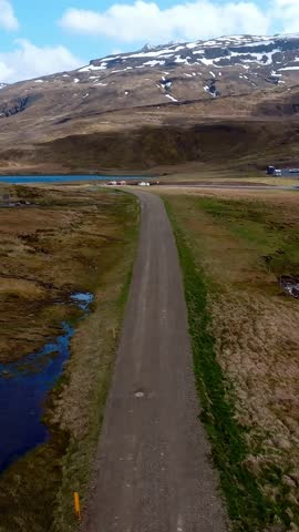 A lone car travels on a dirt road in Iceland, surrounded by open landscapes. Snow capped mountains and a small body of water are visible under a partly cloudy sky.