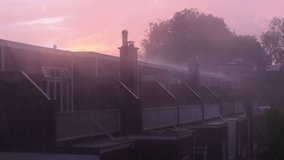 A row of houses with chimneys is seen under a pink-hued sky during rainfall. Raindrops blur the view, and trees in the background add depth to the misty atmosphere. The sunset casts a soft glow. - Powered by Shutterstock - Get 15% off with code: PIKWIZARD15