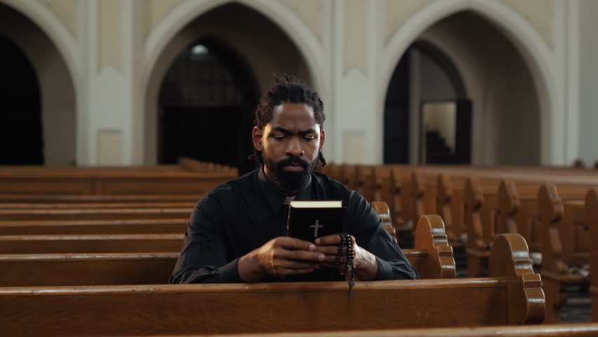 Young priest is sitting in an empty church and praying to god while holding the holy bible and a rosary