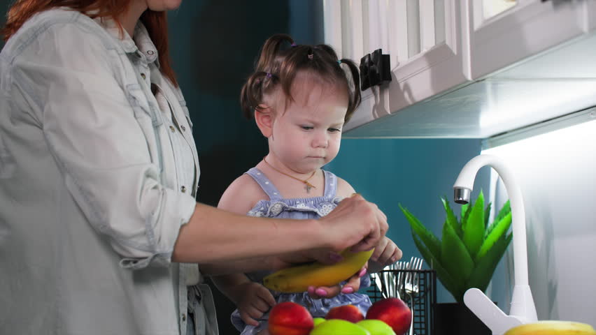 healthy food for kids, caring female parent peeling banana for little cute girl in kitchen