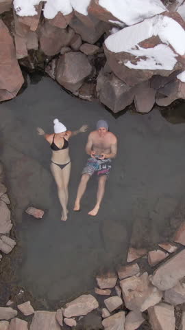 DRONE, TOP DOWN: Tourist couple relaxes in a hot spring in the middle of the snowy wilderness in Colorado. Flying above two young travelers taking a dip in a hot river flowing across wintry Colorado