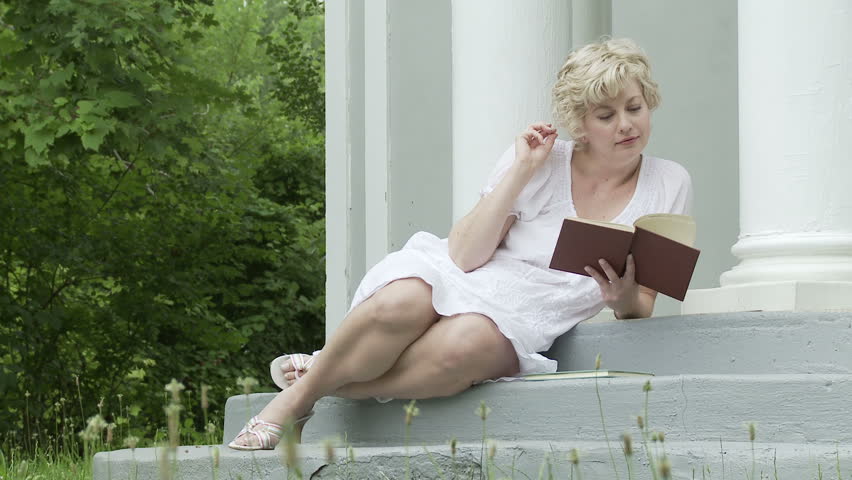 Blonde girl in white dress reading a book, sitting by columns with trees in the