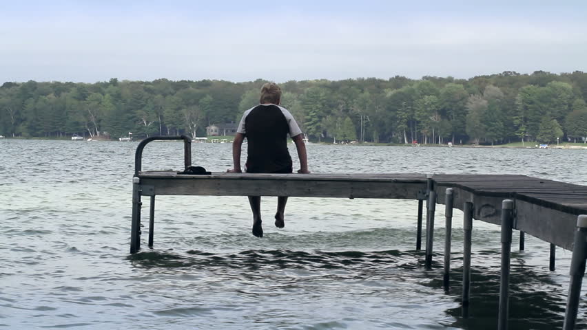 Teenage boy sits on the end of a wooden dock at the side of a lake, splashing in