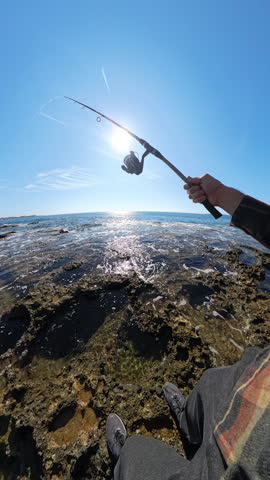 Close first-person view of hands holding a fishing rod while casting on a rocky seashore under a bright blue sky with the sun reflecting on the shimmering ocean water