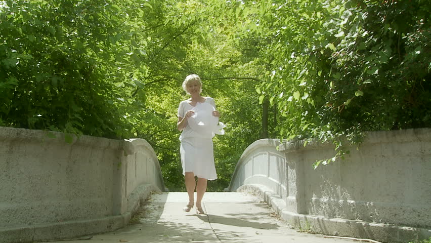 Blonde girl in a white dress and holding a white hat, runs across a bridge
