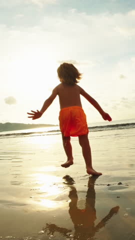 Vertical shot Back view of barefoot kid running on waves and jump in water. Concept of happy childhood, memories of family weekend, trip. Child funny game, plays at sunset, enjoy moment