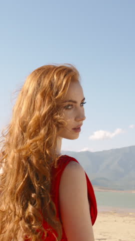 Vertical Video Redhead Female Wearing Red Dress on Beautiful Landscape, 20s Girl Looking at Camera Wind Waves Her Red Dress. Gorgeous Lovely coquettish Woman with Long Curly Red Hair. Freedom concept