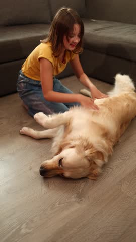 A cheerful teen girl is lying on the floor, playfully petting a golden retriever. Their cozy living room setting creates a warm atmosphere filled with joy and companionship.