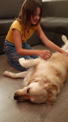 A cheerful teen girl is lying on the floor, playfully petting a golden retriever. Their cozy living room setting creates a warm atmosphere filled with joy and companionship.