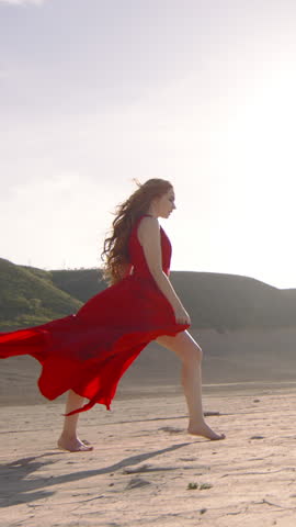 Vertical shot of happy woman running in the desert. Long red silk dress and red hair flying fluttering in the wind in slow motion. A tourist is enjoying a vacation in the Dubai desert