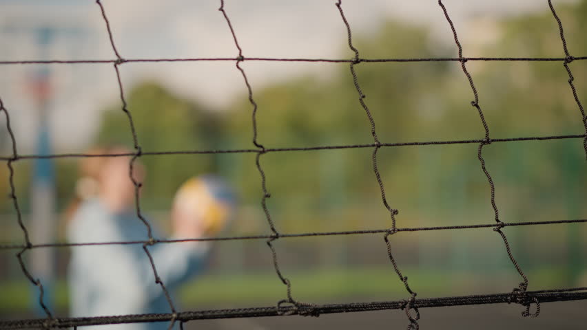 Close-up of volleyball net with blurred background of two people playing volleyball, sunlight creates golden glow on greenery, enhancing energetic outdoor scene of sports activity