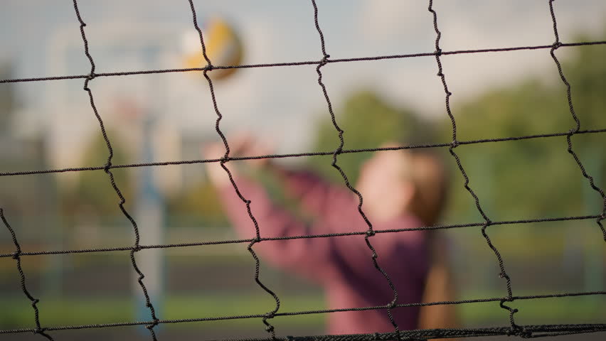 Close-up of volleyball net with blurred background showing lady in maroon playing volleyball with another person, sunlight and greenery create golden glow