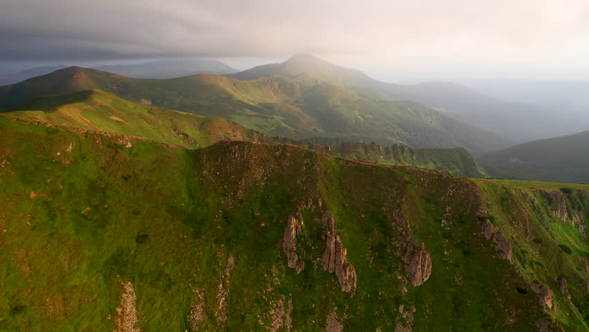 Breathtaking aerial view of green, rugged mountain peaks under dramatic sky. Scene captures contrast between vibrant landscape and moody clouds, with sunlight breaking through. Carpathians, Chornogora