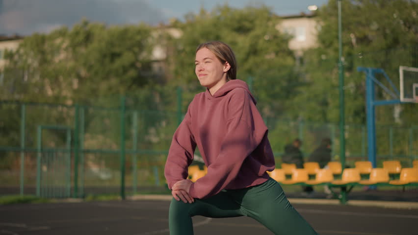 Close-up of lady stretching in green leggings, pink hoodie, outdoor court background with sports equipment and seating area and trees with greenery