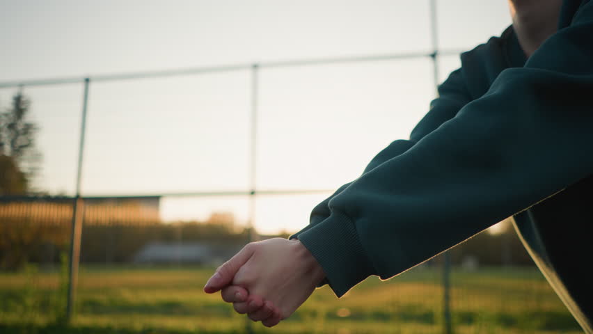 Close-up hand view of someone in green hoodie playing volleyball with sunlight glow effect in background, featuring open field and distant building during golden hour