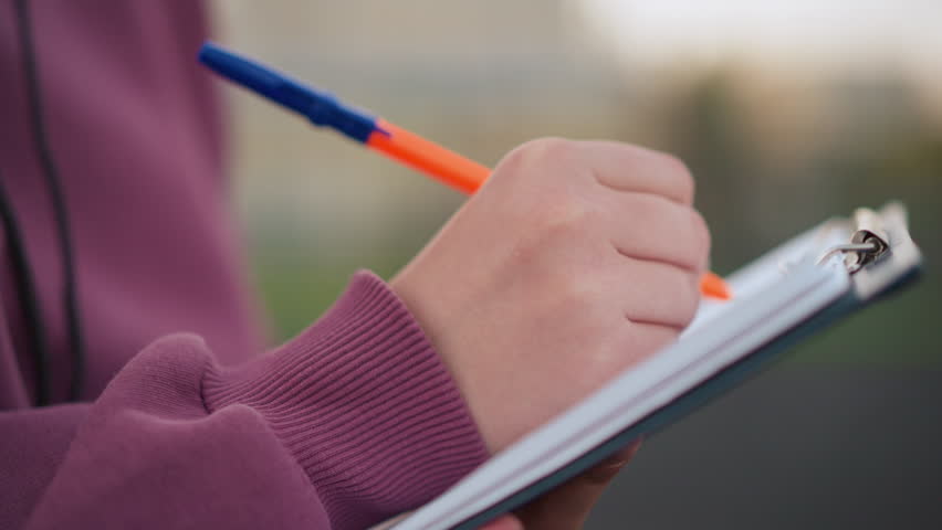 Close-up side view of individual in maroon top taking notes on clipboard outdoors, using an orange pen, background shows sports field with people walking
