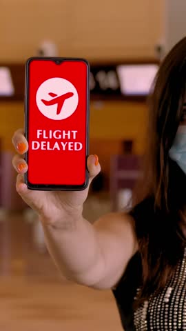 Woman, in protective mask, holds smartphone with airplane icon, text - flight delayed, on screen, in front of empty check-in counters at the airport.