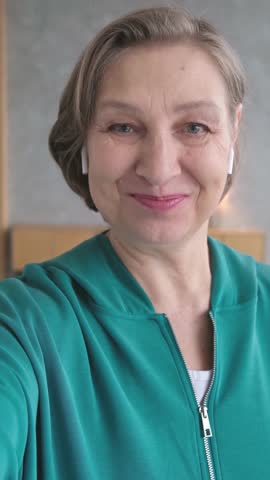 Vetical video of beautiful smiling mature woman with wireless headphones making selfie video call with phone. 