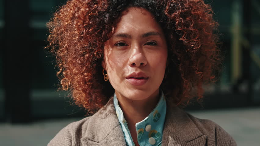 A young businesswoman with curly hair stands in the city, exuding confidence and professionalism. She is ready to take on the day in this urban setting.