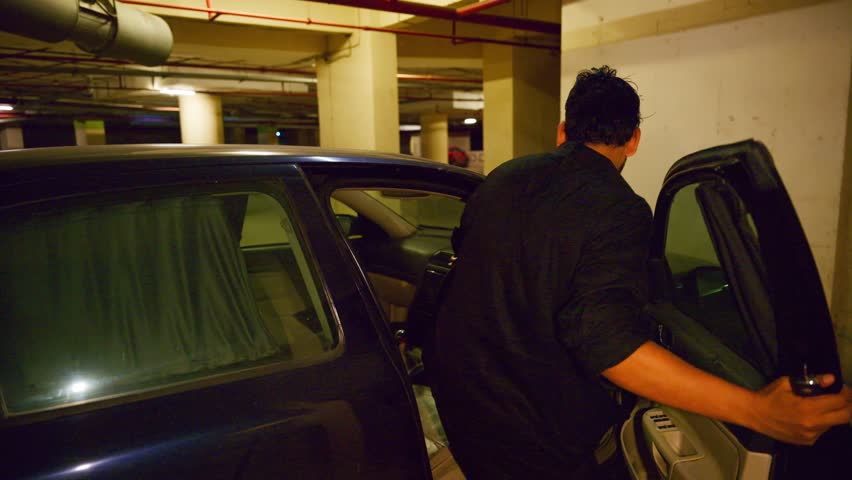 Man Stands Beside Car with Open Door in Basement, Guy Grabs Hat from Dashboard Inside Parked Vehicle, Fellow passes Cap by Throwing It Toward Another, Boy Steps Back Into Motorcar After Passing Object