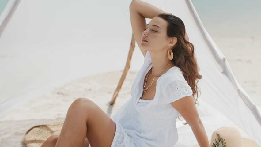 A tanned woman in a boho white dress lounging under a beach tent, surrounded by tropical elements like pineapple and coconut. Her serene pose evokes relaxation, summer vibes, and natural beauty.