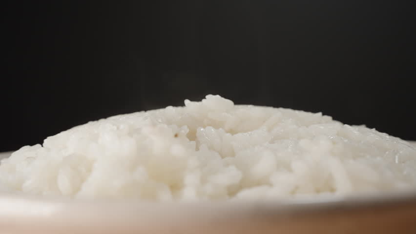 Wooden Spoon Lifting Steaming Freshly Cooked White Rice from a Bowl, Showing Soft Texture and Moisture Rising in Warm Steam Against a Dark Background