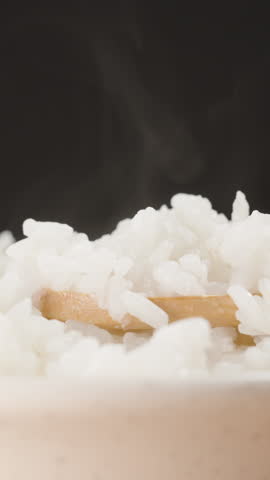 Vertical video. Rising Steam Over a Bowl of Freshly Cooked White Rice as a Wooden Spoon Scoops Up Soft and Moist Grains, Creating a Warm and Comforting Scene