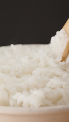 Vertical video. Close-Up of a Hand Using a Wooden Spoon to Scoop Freshly Cooked White Rice from a Steaming Bowl, Emphasizing Softness and Warmth