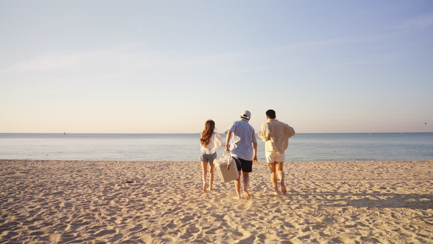 Happy man and woman travel the sea on summer beach holiday vacation. Group of Asian people friends enjoy and fun outdoor lifestyle holding cooler box walking to picnic on tropical beach in sunny day.