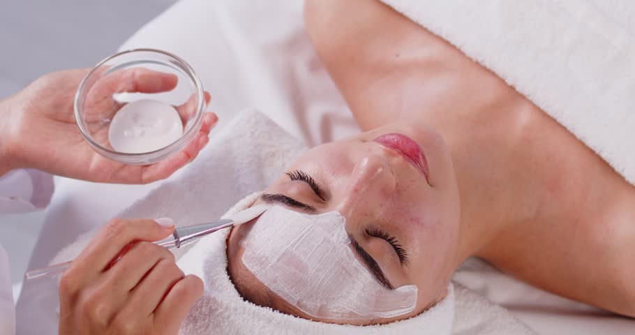 Woman getting peeling spa beauty mask, getting facial care, enhancing her skins radiance, undergoing beauty procedures, lying on couch with wrapped towel on head, enjoying moment of relaxation