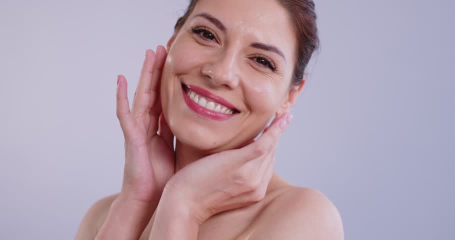 Slow motion video of a young beauty laughing woman touching her healthy facial skin and looking cheerful at camera isolated on a gray background. Skin and body care concept. 4k video.