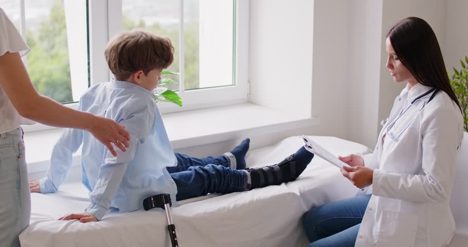 Little boy visiting pediatrician doctor on couch, friendly female paediatrician exams leg, symptoms, talking to kid parent, traumatologist physician provides consultation, healthcare, medical check-up
