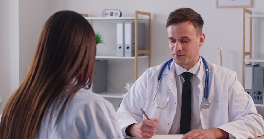 Male GP doctor giving consultation to patient in hospital office, listening to complaints, writing medical records, dosage in register, woman visiting, employee consulting therapist to get sick leave