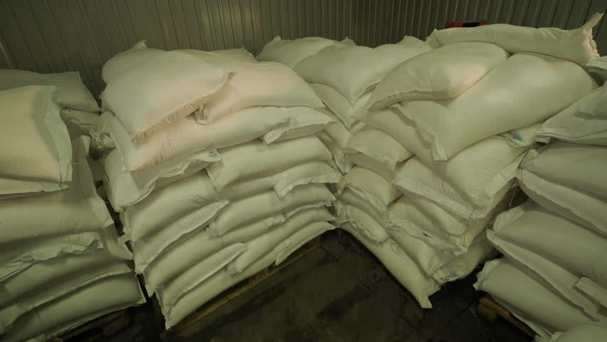 Large stacks of white sugar bags stored in a warehouse, organized on pallets under artificial lighting in an industrial storage facility.